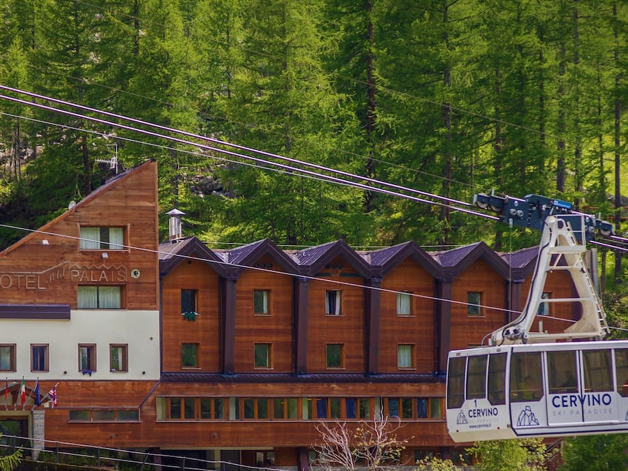 A wooden building with a sign that reads "HOTEL PALAISE" and a cable car labeled "CERVINO" in front of it.
