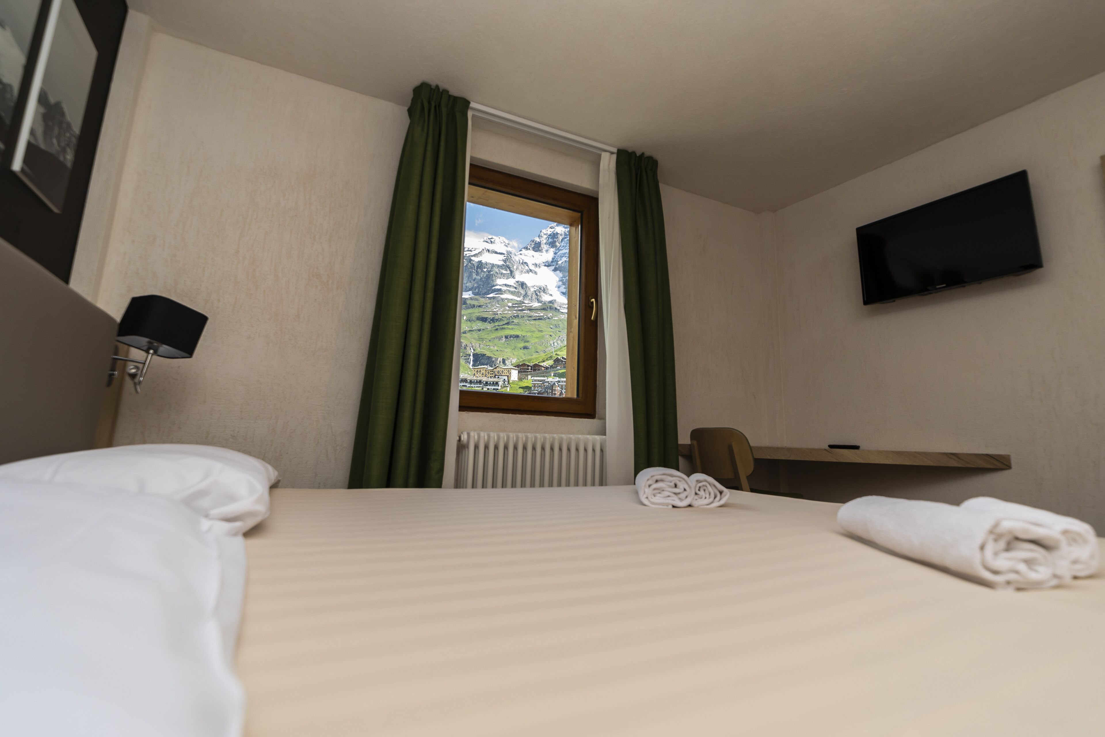 A hotel room with a bed, a window with curtains, a TV mounted on the wall, and a radiator.