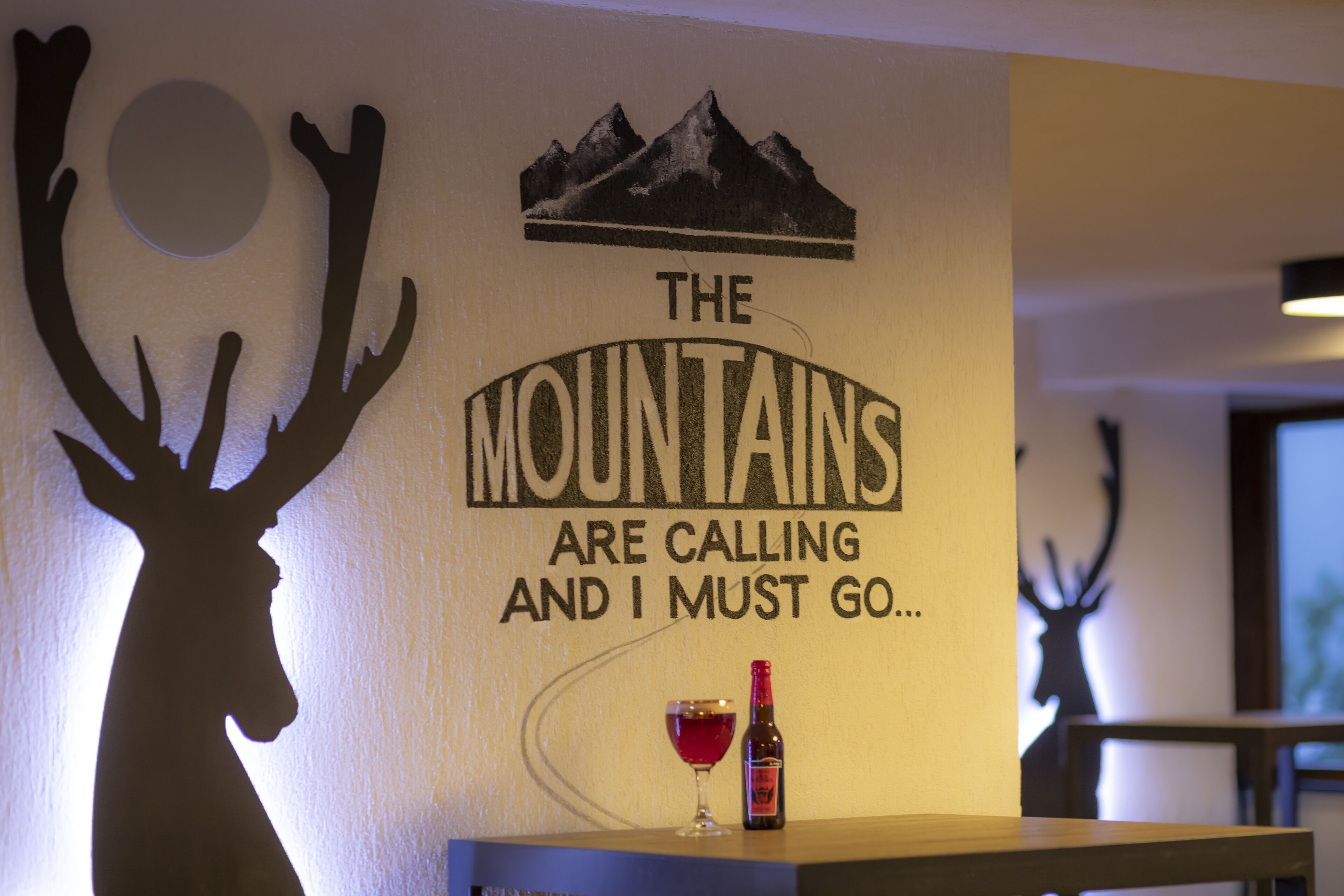 Una pared con la silueta de una montaña y texto, la silueta de un ciervo y una botella de vino sobre una mesa.