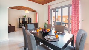 A dining area with a dark wooden table set for four, surrounded by grey chairs. A fruit bowl with grapes and oranges is on the table, and there are glasses of orange juice. The room has a modern design with a wooden ceiling and a large window with red patterned curtains. - Villa Vista Mar - Holiday Home (Funchal)