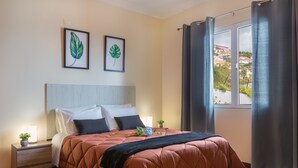 A neatly made bed with a brown quilt, two framed leaf artworks on the wall, a bedside table with a lamp, and a window with curtains. - Villa Vista Mar - Holiday Home (Funchal)