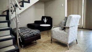 Una sala de estar moderna con una escalera, un sofá negro, un otoman estampado, un sillón blanco y una pequeña mesa redonda con un vaso.