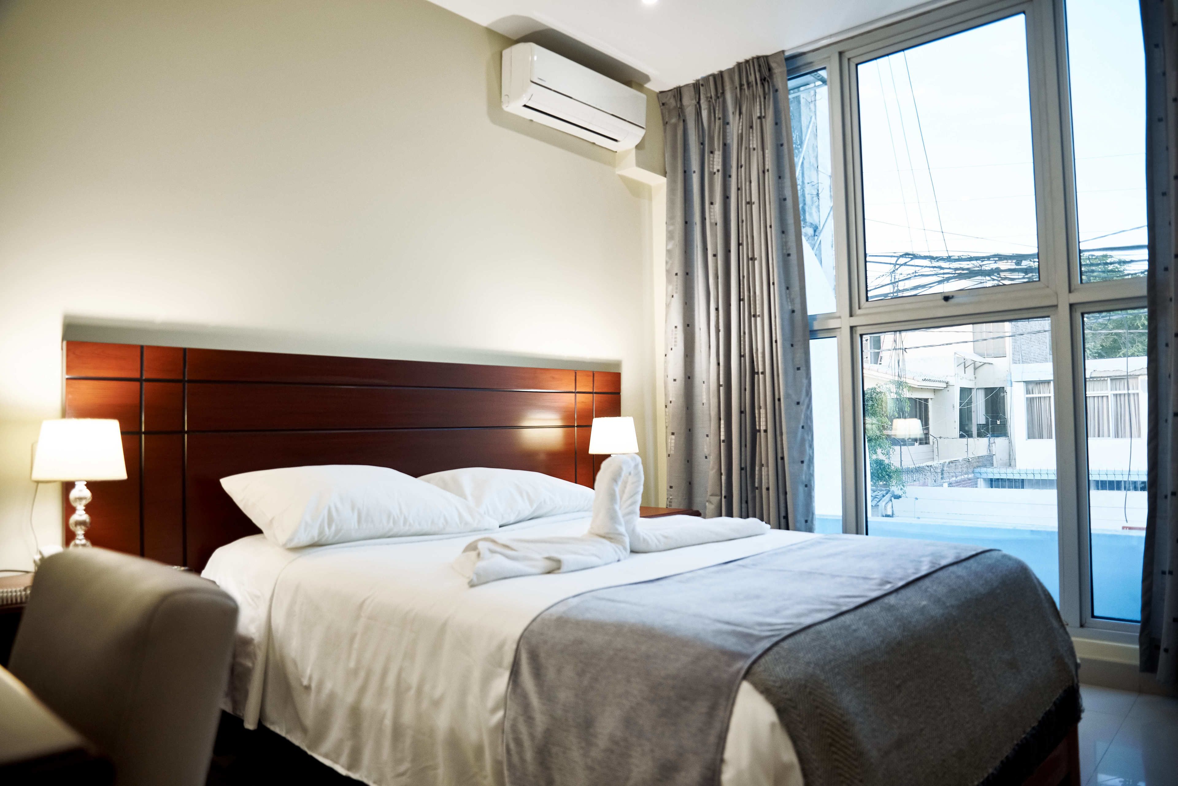 Una habitación de hotel con una cama grande, un cabecero de madera, una ventana con cortinas y un equipo de aire acondicionado.
