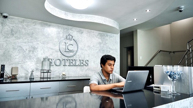 Una recepción de hotel con una pared de mármol, un logotipo con forma de corona y un ordenador portátil sobre el mostrador.