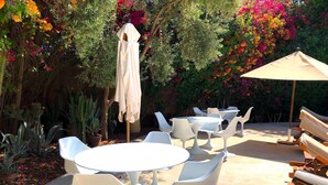 Un patio al aire libre con mesas y sillas blancas, rodeado de exuberante vegetación y coloridas flores.