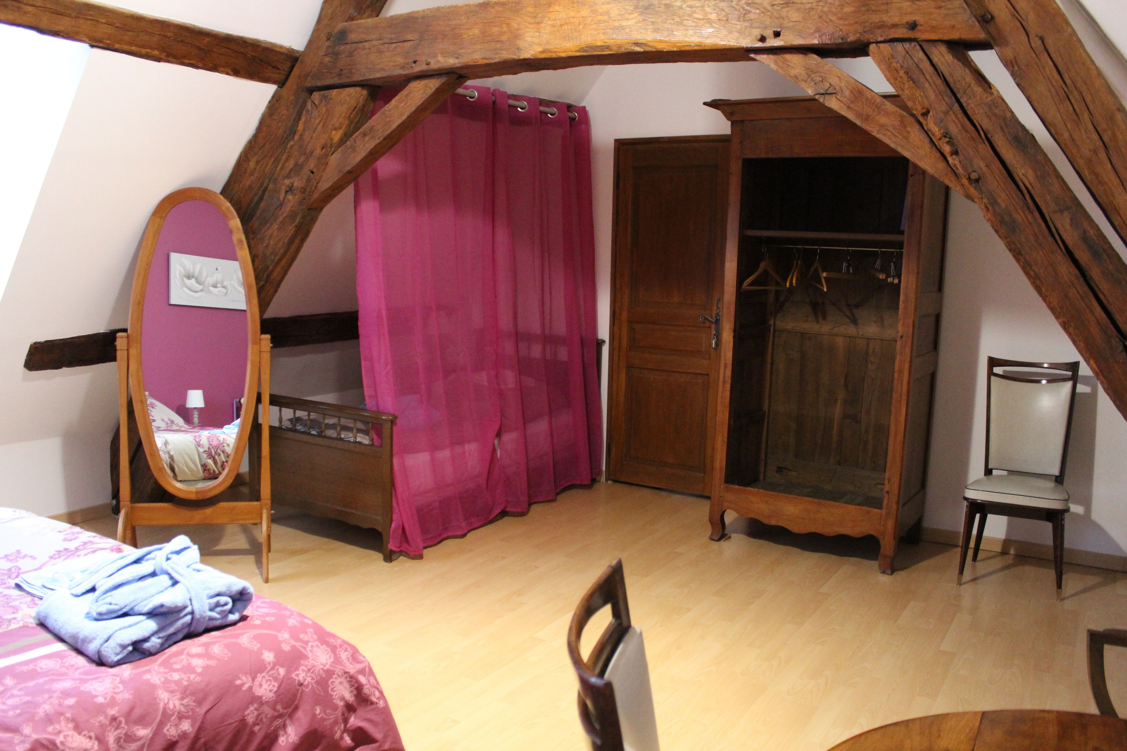Een slaapkamer met een houten plafond, een bed met een roze dekbedovertrek, een houten garderobe, een stoel en een houten kast met daarin hangende kleding.