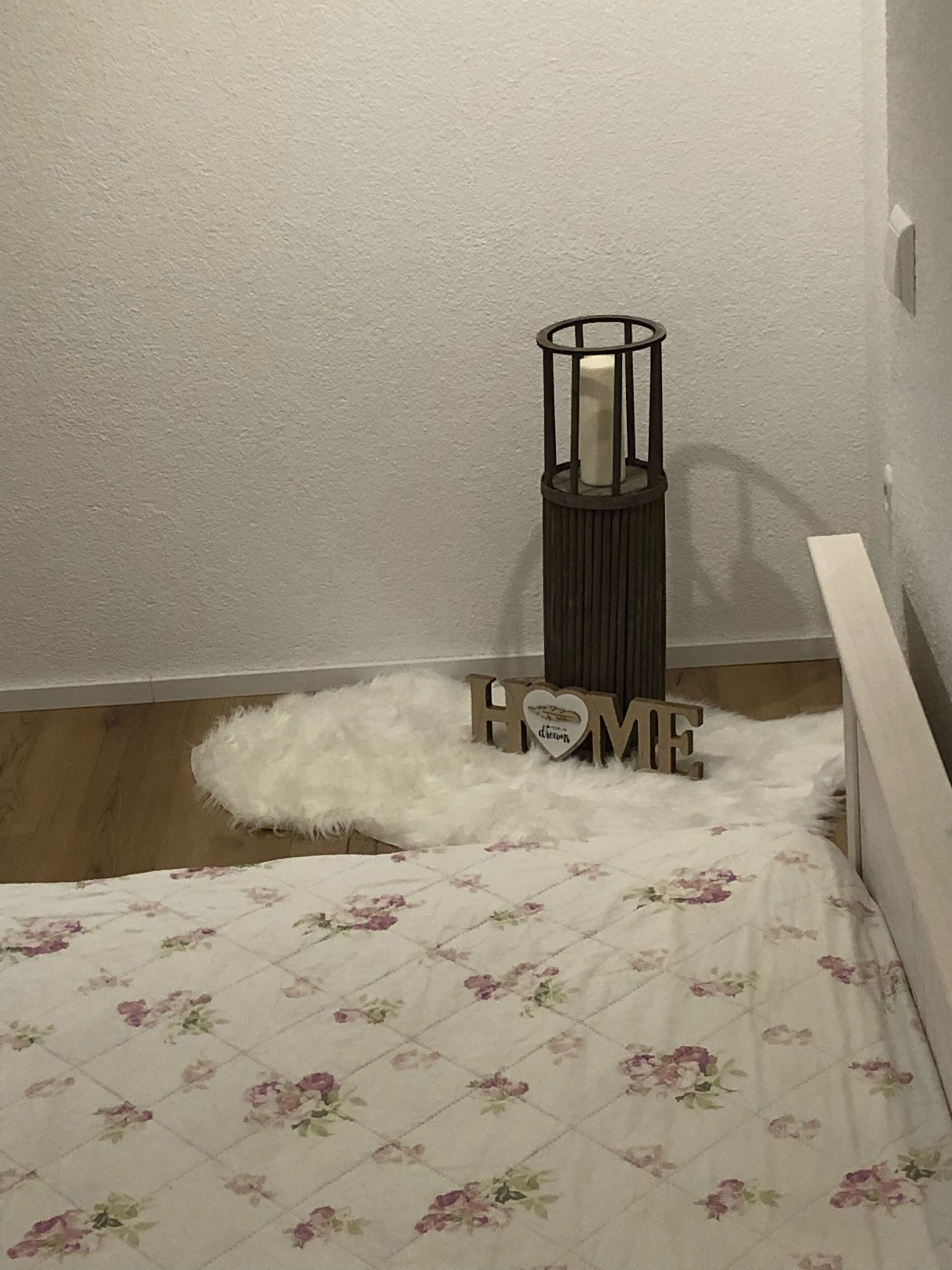 Et soveværelse med en seng, en blød hvid måtte, en vægmonteret lysestage og et skilt med teksten 'HOME'.