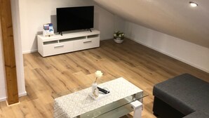 Un salon moderne avec un téléviseur à écran plat posé sur un meuble blanc, une table basse en verre agrémentée d’un objet décoratif, un canapé gris et un plancher en bois.