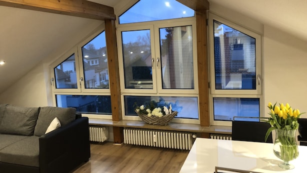 Une pièce de style loft avec de grandes fenêtres, un canapé noir, une table blanche et un vase rempli de fleurs jaunes.