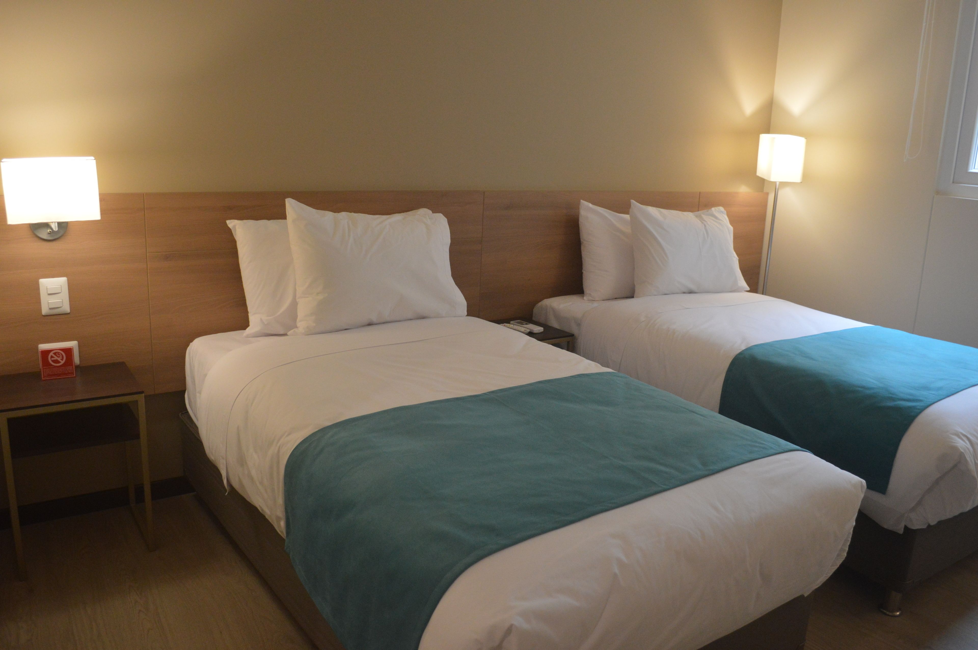 A hotel room with two beds, each with white bedding and teal throw pillows. There are bedside lamps on either side of the beds, and a small table with a red sign between the beds.