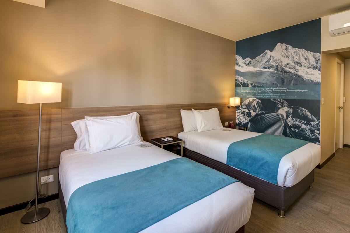 A hotel room with two beds, a nightstand, a lamp, and a large wall mural of a mountain landscape.