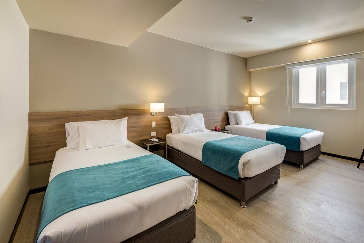 A hotel room with two beds, each with a wooden headboard, white bedding, and teal throw blankets. There are bedside tables with lamps and a window with blinds.