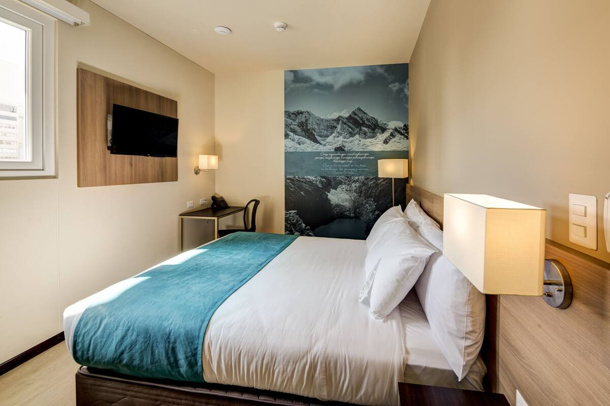 A hotel room with a bed, a mounted TV, a desk, a chair, a wall-mounted lamp, and a poster of mountains.