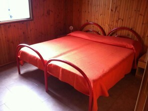 Uma cama de solteiro com estrutura de metal vermelha e roupa de cama vermelha combinando, num quarto com paredes de madeira e uma janela.