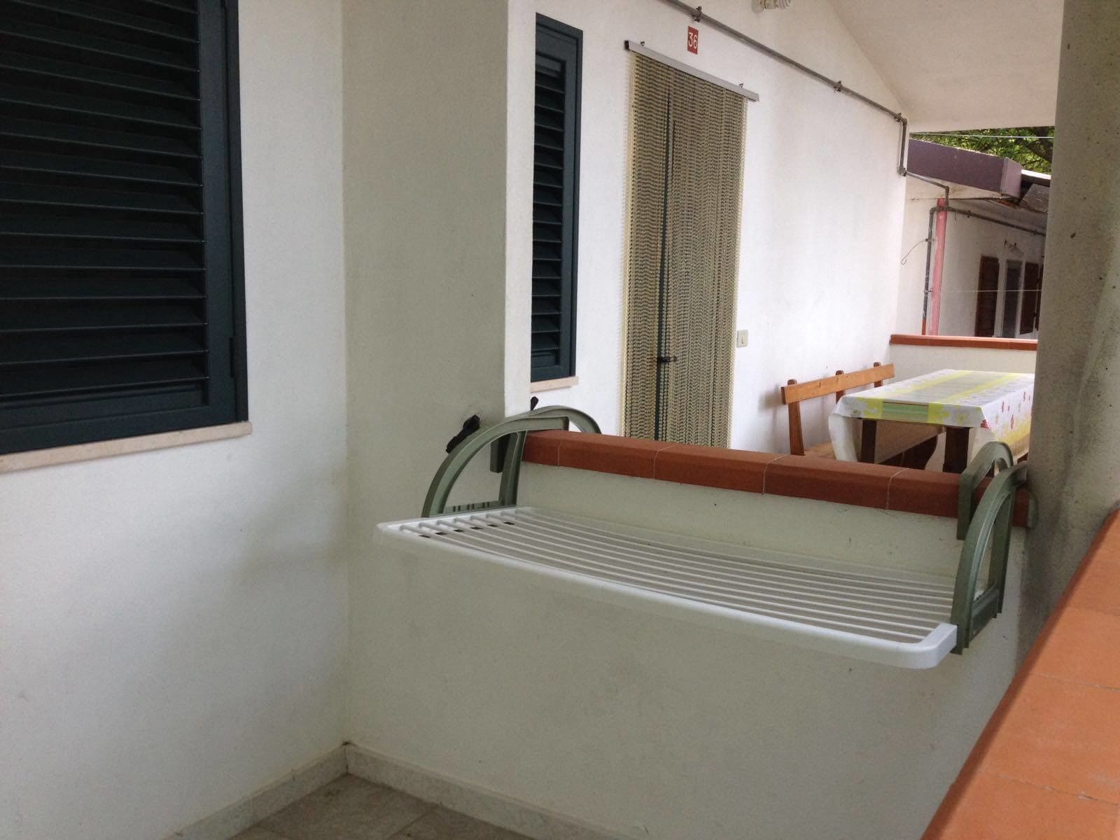 Un piccolo balcone con un portascope in metallo, un tavolo con sedie e una porta con finestra verticale.