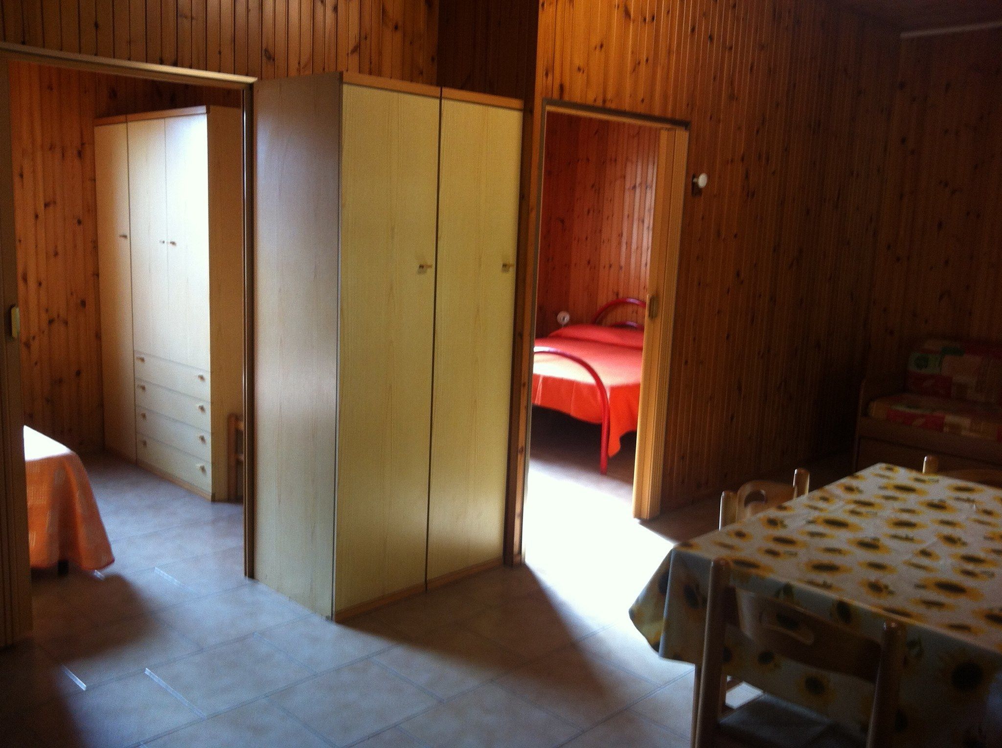 A room with wooden walls, a bed with a red bedspread, a wardrobe, and a table with a patterned tablecloth.