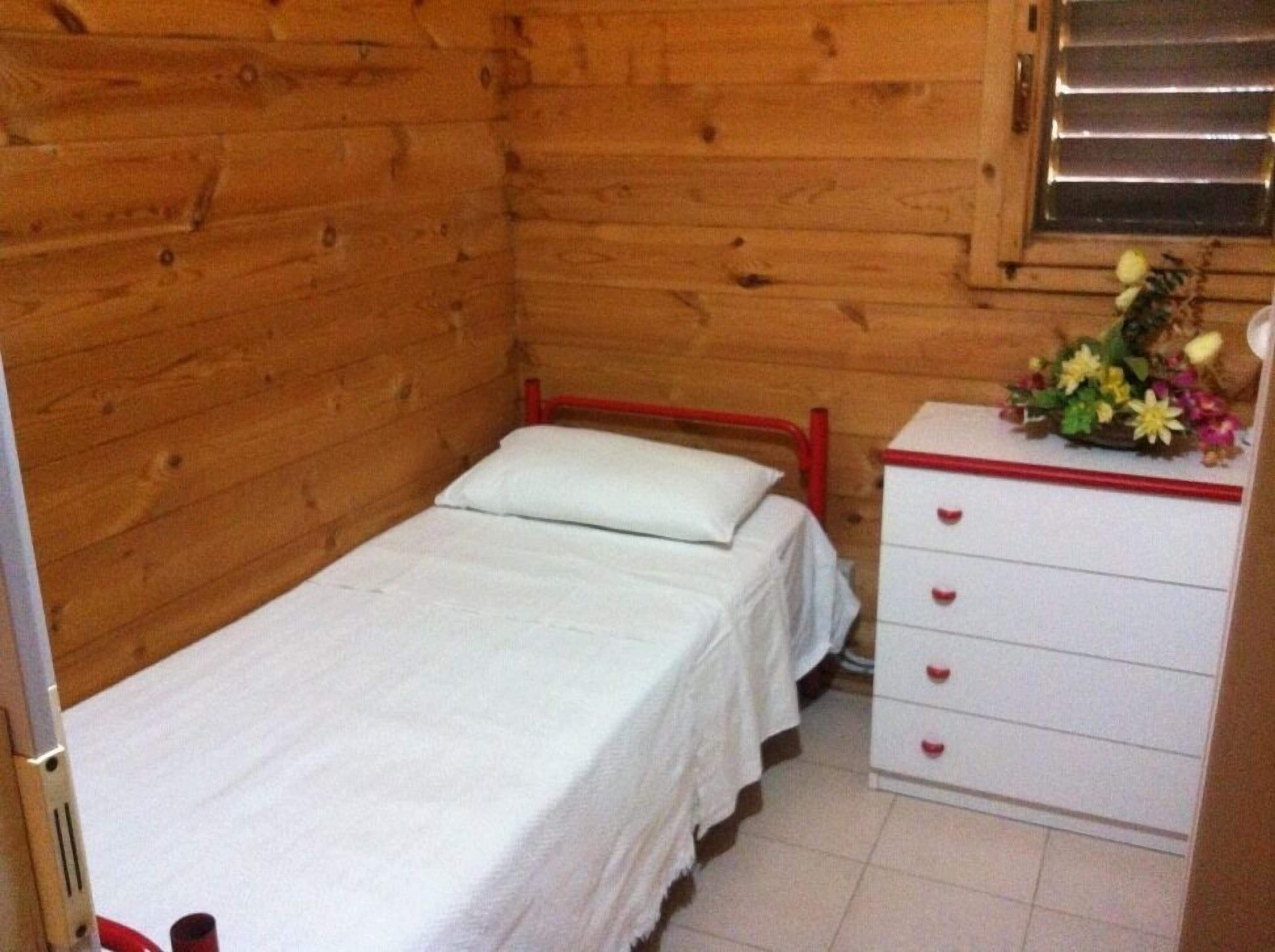 Une petite pièce en bois avec un lit simple, une commode à tiroirs et une fenêtre avec des stores.
