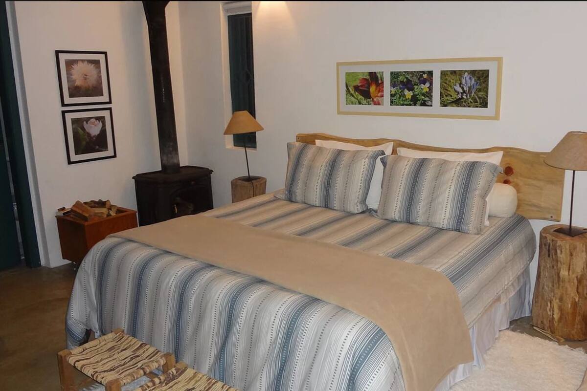 A bedroom with a large bed, two bedside tables, a wooden nightstand, a wall-mounted lamp, and framed pictures on the wall.