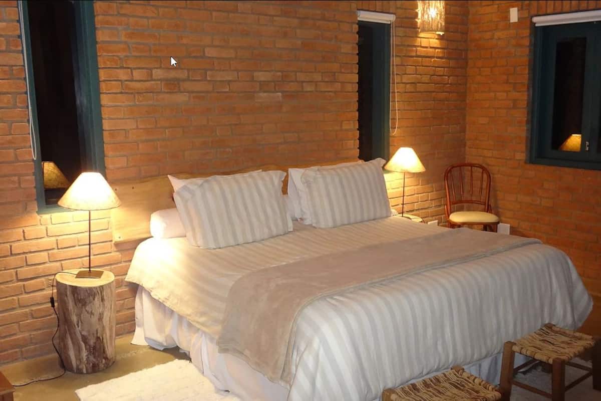 A bedroom with a brick wall, a bed with white bedding, two bedside lamps, and a window with a view.