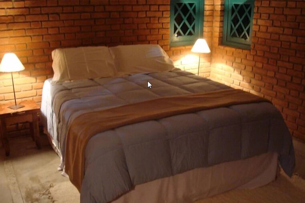 A bedroom with a brick wall, a bed with a grey and white duvet, two bedside lamps, and a wooden nightstand.