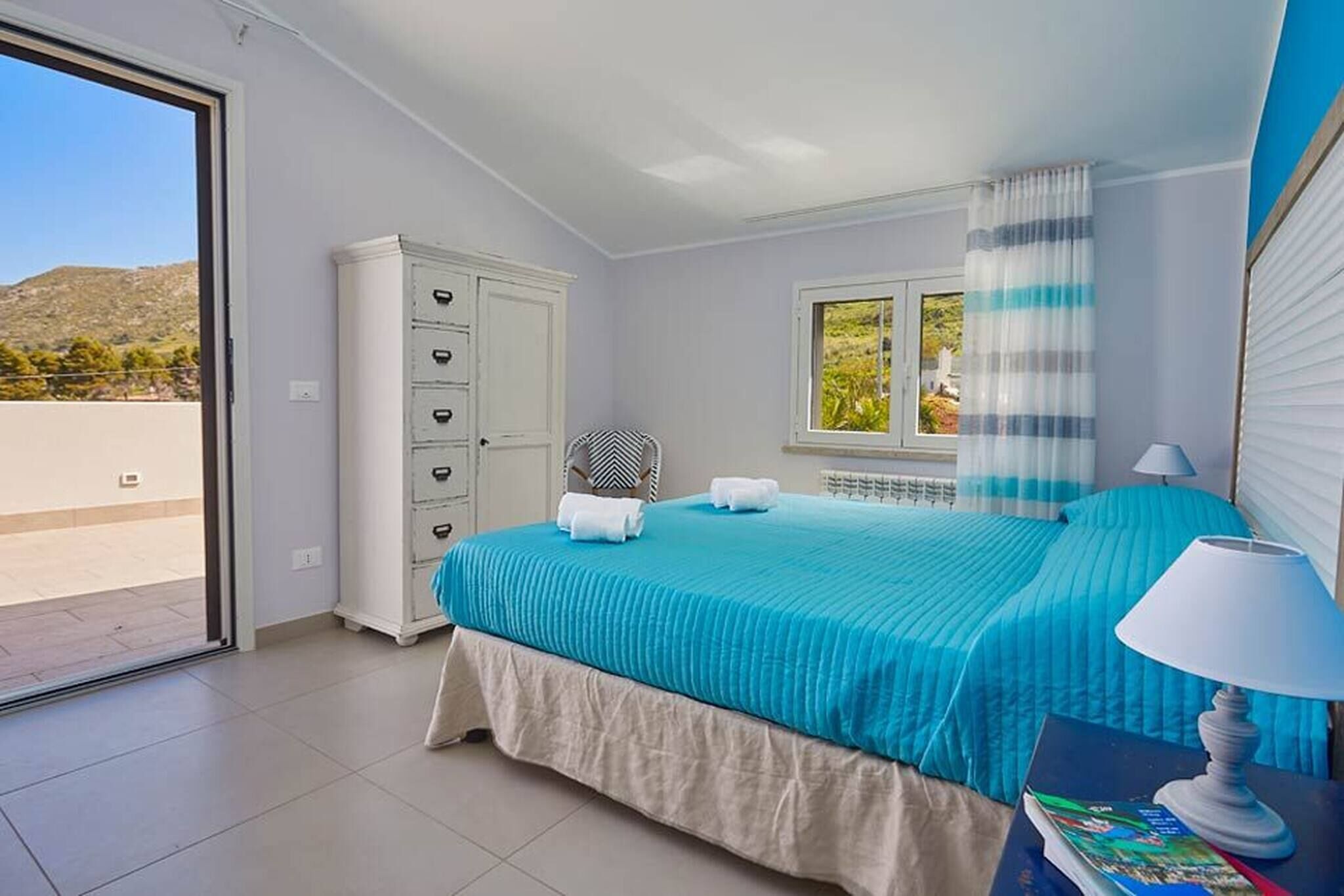 Une chambre à coucher avec un lit, une table de chevet avec une lampe, une fenêtre avec des rideaux et une armoire blanche.