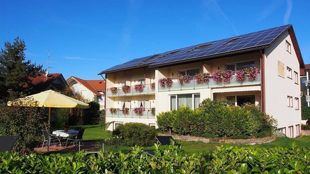 Ein zweistöckiges Gebäude mit Solaranlage auf dem Dach, Balkonen mit Blumenkästen und einer Terrasse mit Tisch und Stühlen.