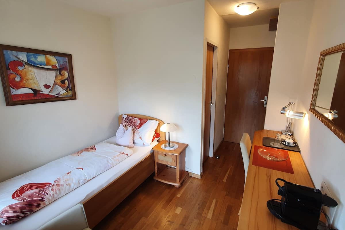 Une petite chambre à coucher, bien rangée, avec un lit simple, une table de chevet avec une lampe, un miroir et un bureau en bois avec une chaise.