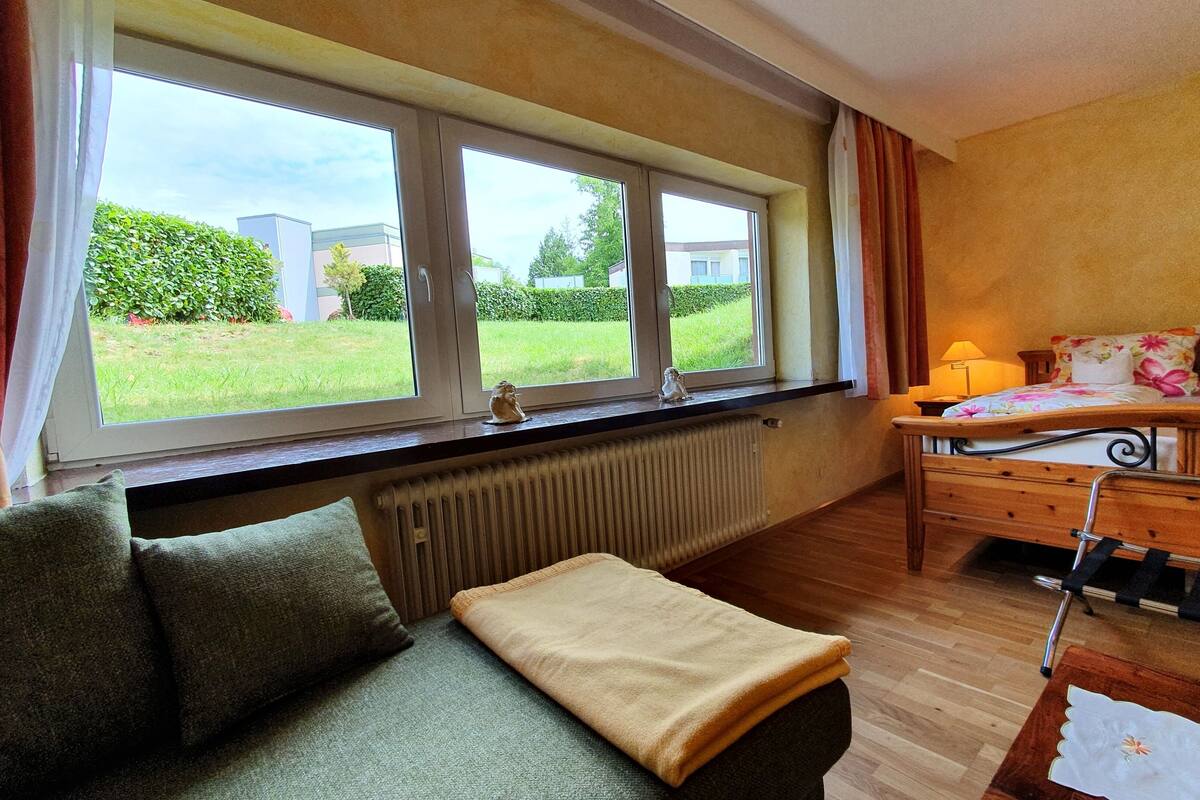 Une chambre à coucher avec un lit, une fenêtre avec des rideaux, un radiateur, un canapé et une vue sur l’extérieur.