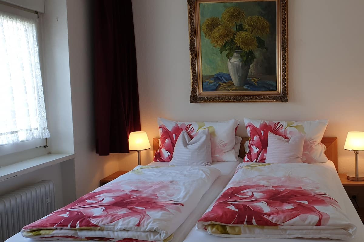 Un lit double avec une literie à motifs floraux, deux lampes de chevet, un tableau au mur et une fenêtre avec des rideaux.