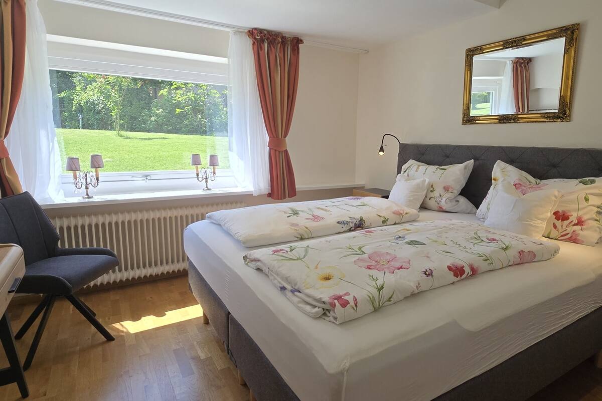 Une chambre avec un lit, un bureau et une chaise. Il y a une fenêtre avec des rideaux, un radiateur et un miroir au mur.