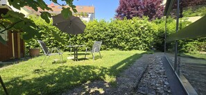 A patio area with a table and chairs, a large umbrella, and a view of a garden and buildings in the background. - Casa Cristina- Kurpension und Appartements (Bad Krozingen)