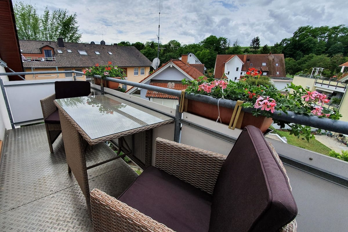 Un balcon doté d’une table en verre et de chaises en osier, offrant une vue sur un quartier résidentiel avec ses maisons et sa verdure.