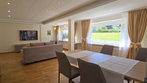 Ein modernes Wohnzimmer mit einer Couch, einem Esstisch und Stühlen. Ein Fernseher ist an der Wand befestigt und es gibt ein Fenster mit Vorhängen.