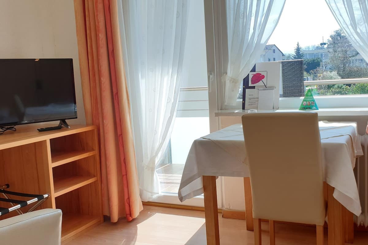Une pièce avec un téléviseur posé sur un meuble en bois, une table recouverte d’une nappe blanche, une chaise et une fenêtre avec des rideaux.