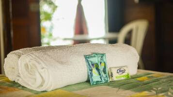 Une serviette blanche, soigneusement pliée, est posée sur un lit recouvert d’une couverture à motifs verts et jaunes. Deux petits emballages de produits de toilette se trouvent à côté de la serviette.