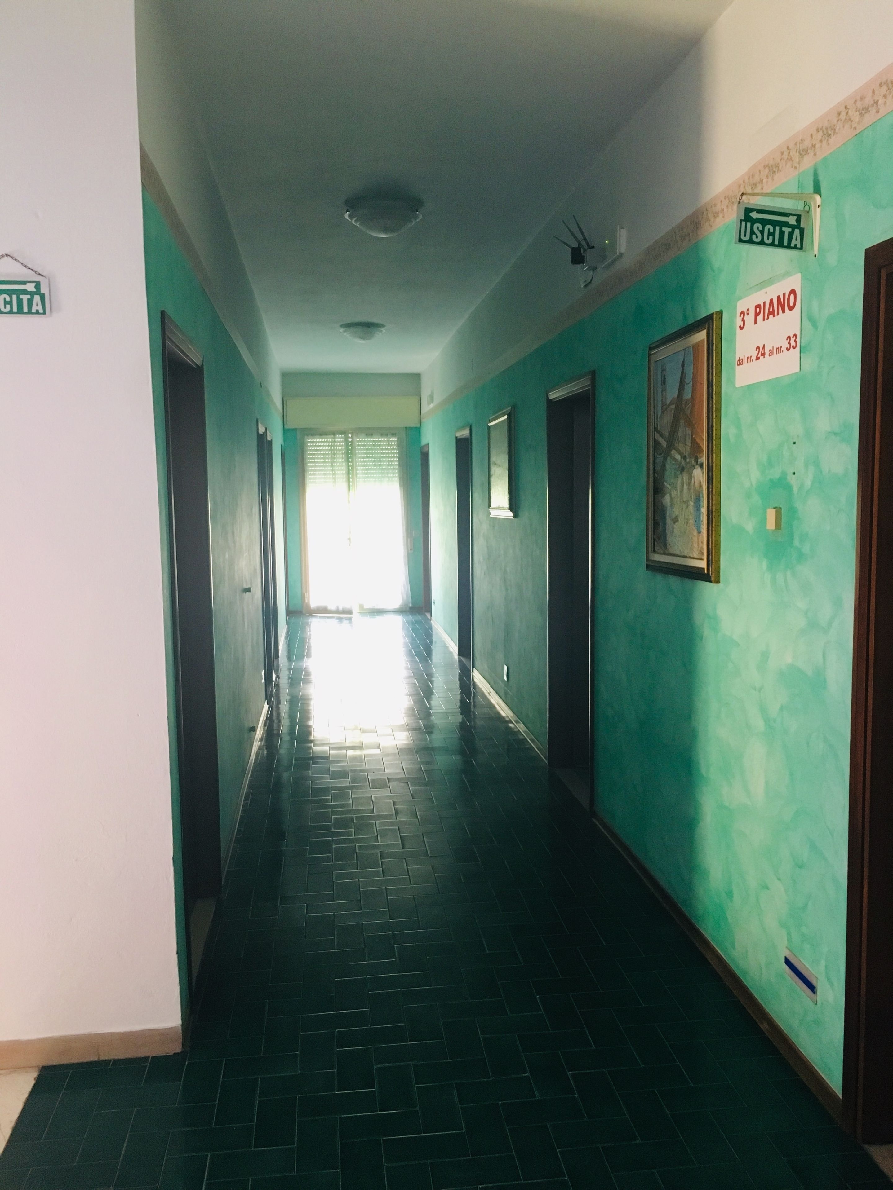 A long, narrow hallway with green walls and black tiled floors. There are doors on both sides, and a sign on the right wall indicates a piano in room 24 and 33.