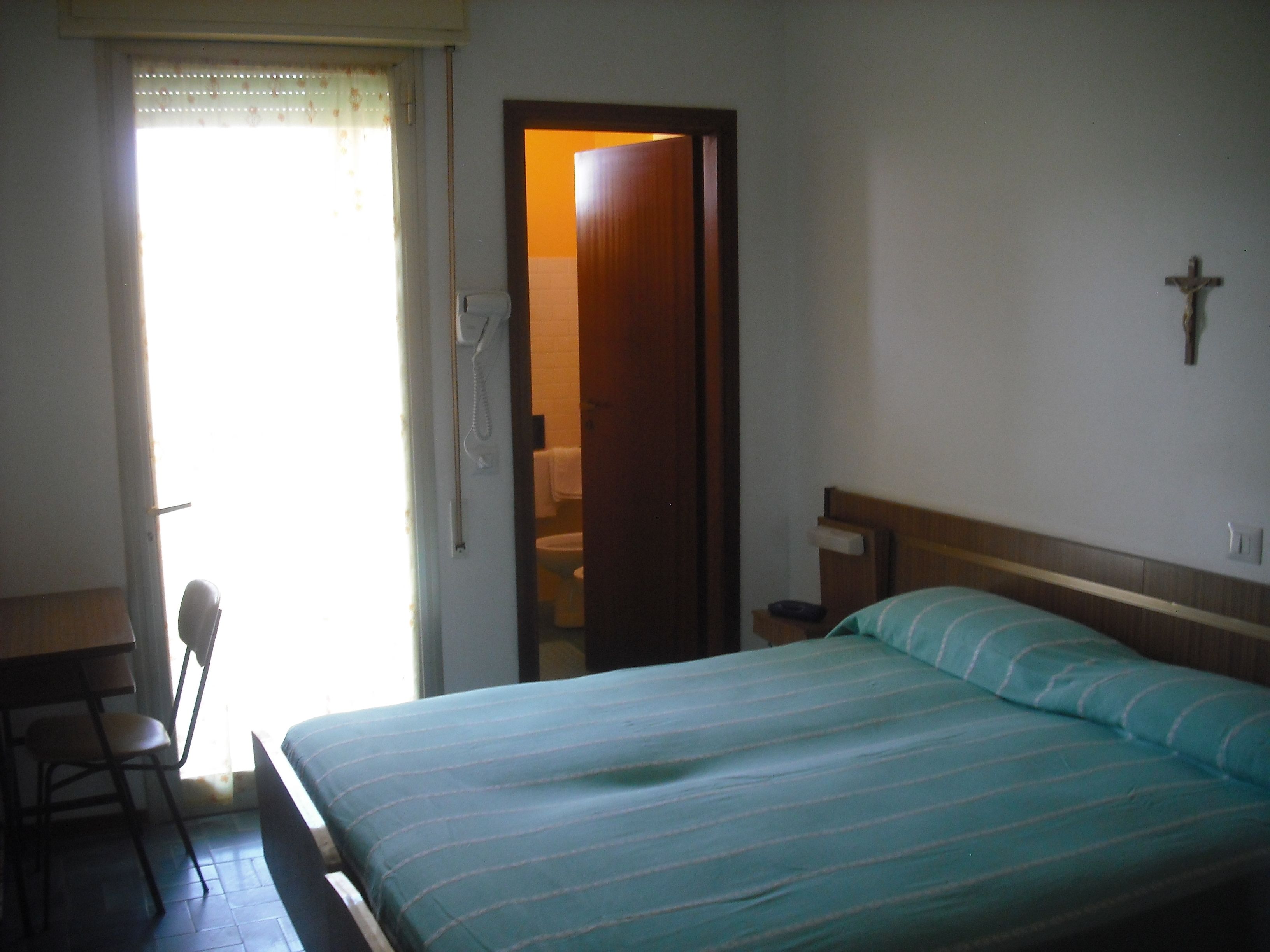 Una camera da letto con un letto, una scrivania e una sedia. Si vede una croce appesa al muro e una porta che dà su un'altra stanza.