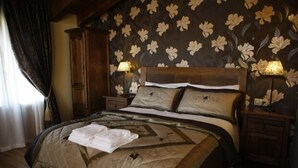 Un dormitorio con una cama grande, papel tapiz floral, cabecera de madera, dos mesitas de noche con lámparas y una ventana con cortinas.