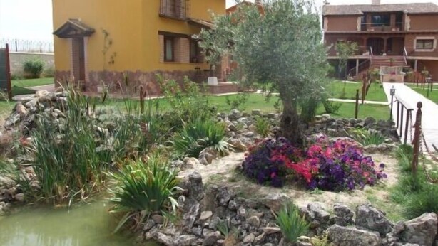 Un edificio amarillo con balcón, rodeado de un jardín ajardinado con estanque y flores de colores.