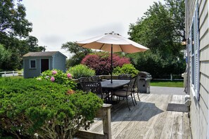 Eine Holzdielen-Terrasse mit Tisch und Stühlen, umgeben von üppigem Grün und einem kleinen Schuppen.