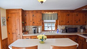 Una cocina con armarios de madera, encimera blanca, un lavamanos, microondas y una mesa con sillas.