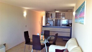 Een woonkamer met een witte bank, een glazen eettafel met vier stoelen, en een keuken op de achtergrond.