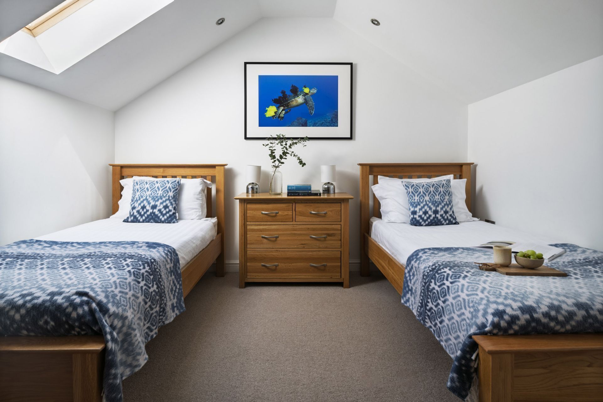 A bedroom with two beds, a nightstand, a framed picture, and a skylight.