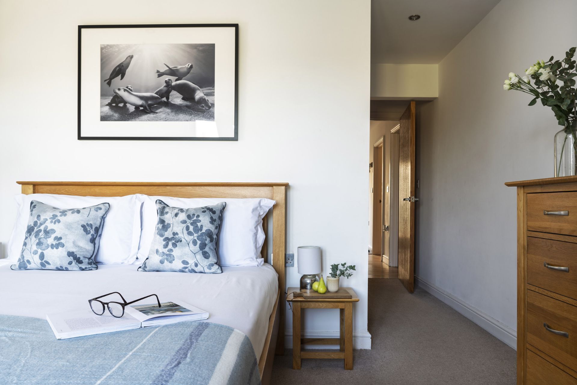 A bedroom with a bed, pillows, a bedside table with a lamp, a framed picture on the wall, and a dresser.
