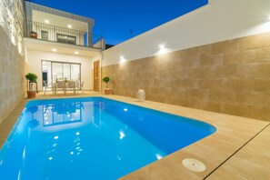 A modern outdoor swimming pool with a clear blue water surface, surrounded by beige tiled walls and a balcony with outdoor furniture.