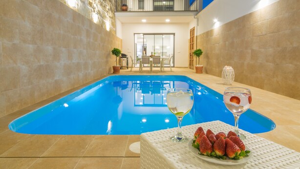 A swimming pool with a white table and chairs, two potted plants, and a glass of water with a strawberry garnish.
