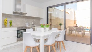 A modern kitchen with white cabinets, a stainless steel range hood, and a built-in oven. There is a dining table with white chairs and a potted plant.