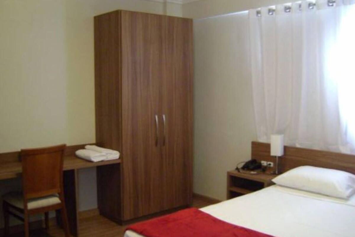 A hotel room with a wooden wardrobe, a desk with a chair, a bed with a red blanket, a nightstand with a lamp, and a window with curtains.