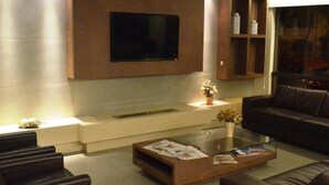 Una sala de estar moderna con un televisor de pantalla plana montado en la pared, un sofá de cuero oscuro, una mesa de centro de vidrio con revistas y una pequeña planta sobre una mesita auxiliar.