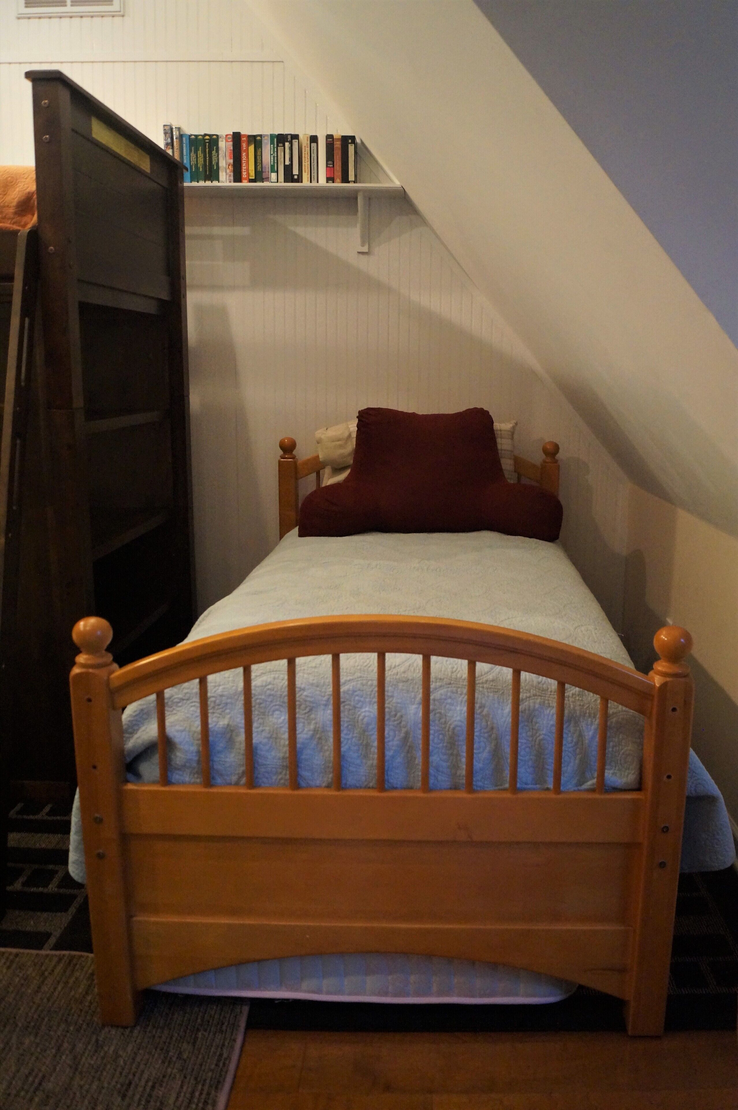 A small, single-bed room with a sloped ceiling, a wooden headboard, a red pillow, and a bookshelf with books.